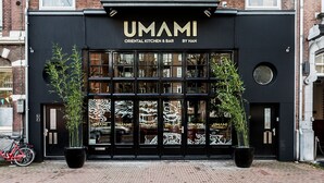 A two-story building with a black facade and white window frames. The ground floor has a black awning with the name "UMAMI" and "ORIENTAL KITCHEN & BAR BY HAN" written on it. There are two potted plants in front of the entrance. - Hotel Iron Horse (Amsterdam)