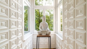 A statue of Buddha on a wooden platform in a room with white patterned walls and a large window. - Home ApartHotel (Kraków)