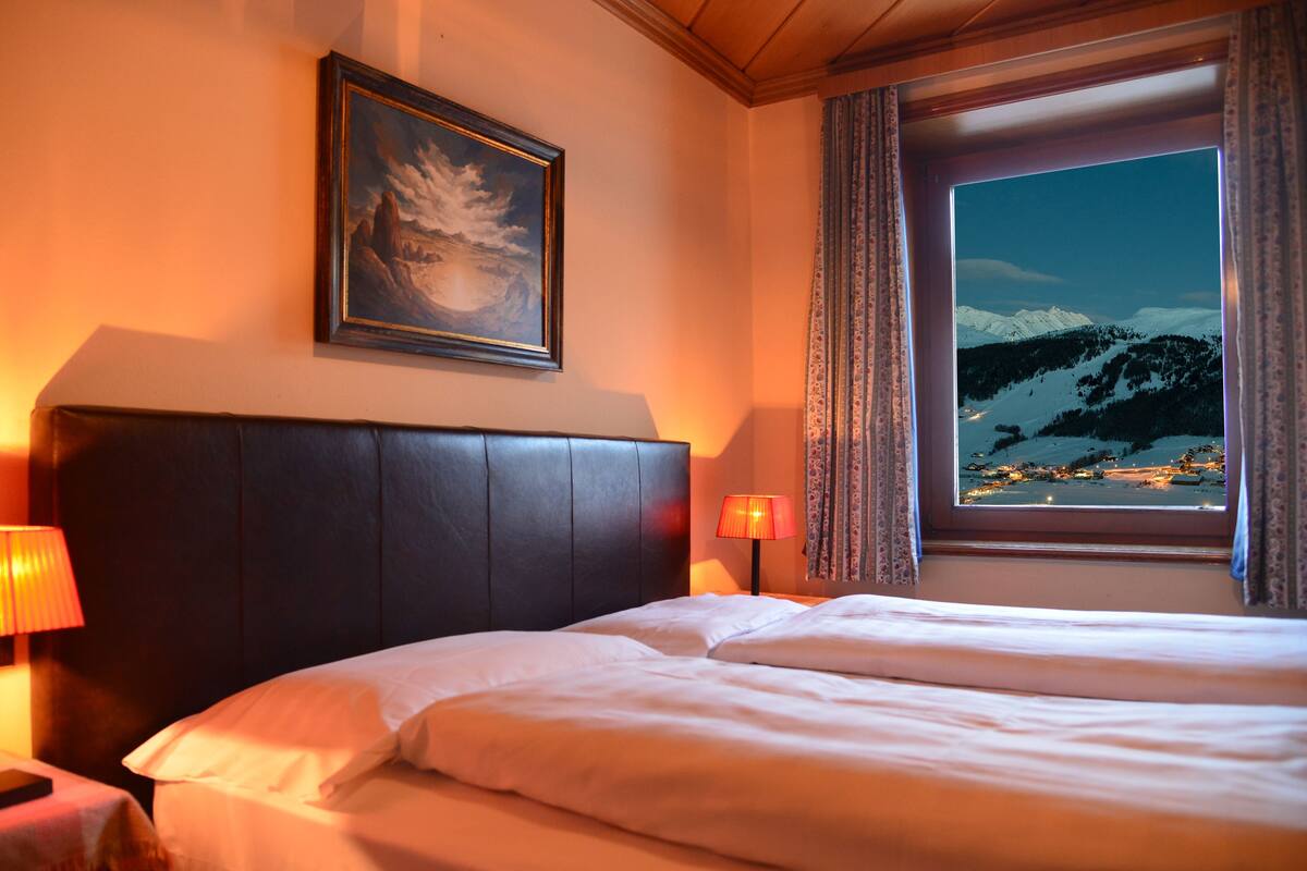 Una camera d'hotel con un letto, un comodino con una lampada, un quadro appeso alla parete e una finestra che si affaccia su montagne innevate.