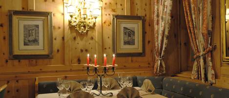 Uma sala de jantar com parede revestida em madeira, quadros, um candelabro com velas e uma cortina floral.