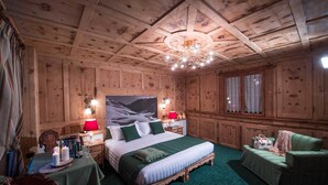 A cozy wooden room with a large bed, two bedside tables with lamps, a green armchair, and a window with curtains. - Alpina (Livigno)