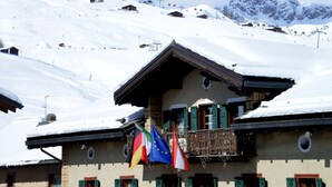 A snow-covered building with multiple balconies, flags, and a mountainous backdrop. - Alpina (Livigno)