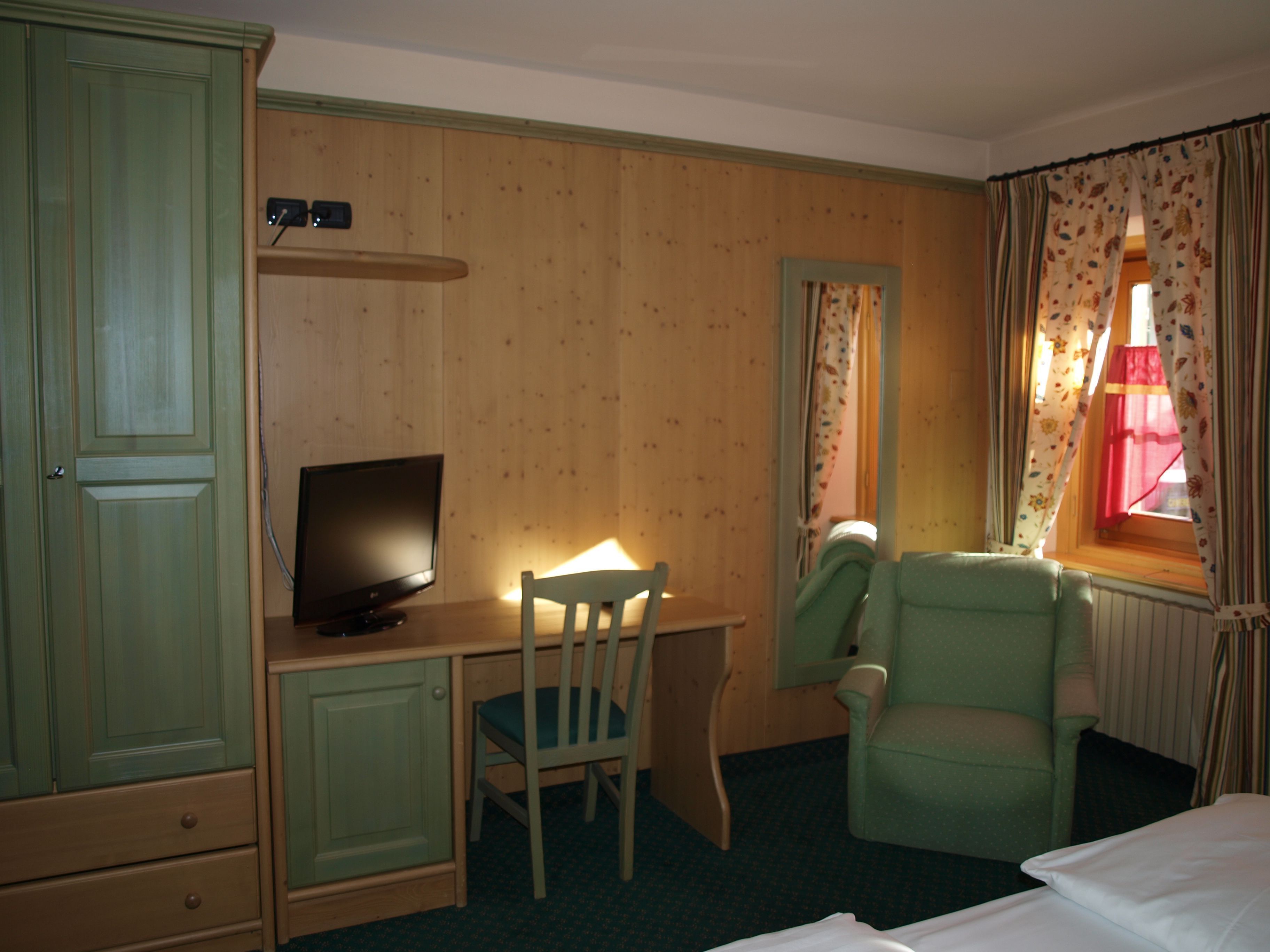 A hotel room with a wooden desk, a television, a green armchair, and a window with curtains.