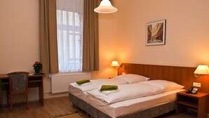 Hotellihuone, jossa on kaksi sänkyä, työpöytä, tuoli, patteri, ikkuna verhoilla ja seinään kiinnitetty lamppu.