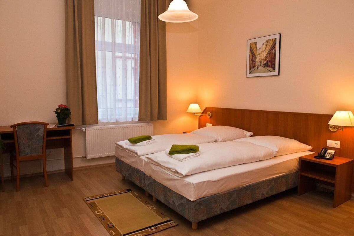 Habitación de hotel con dos camas, un escritorio, una silla, un radiador, una ventana con cortinas y una lámpara de pared.