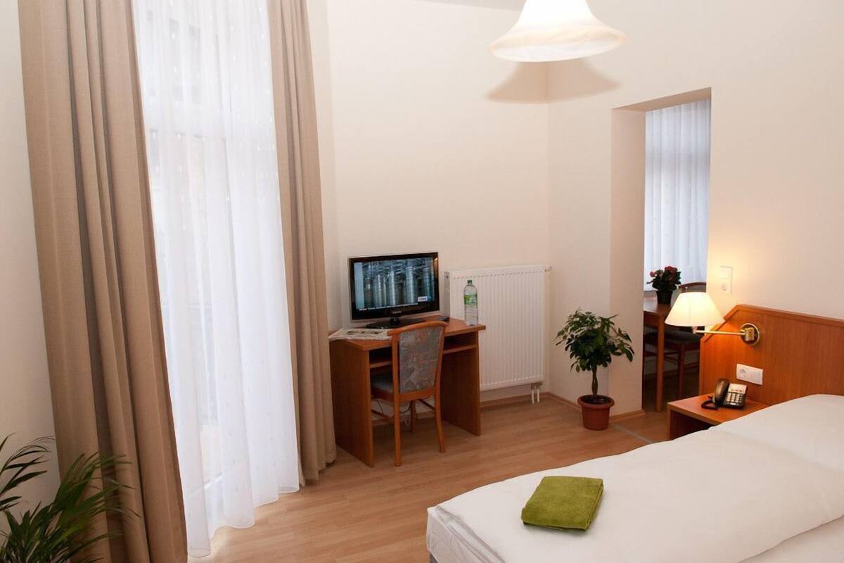 Una habitación de hotel con una cama, un escritorio con una computadora, una silla, una mesita de noche con una lámpara y una planta en maceta.