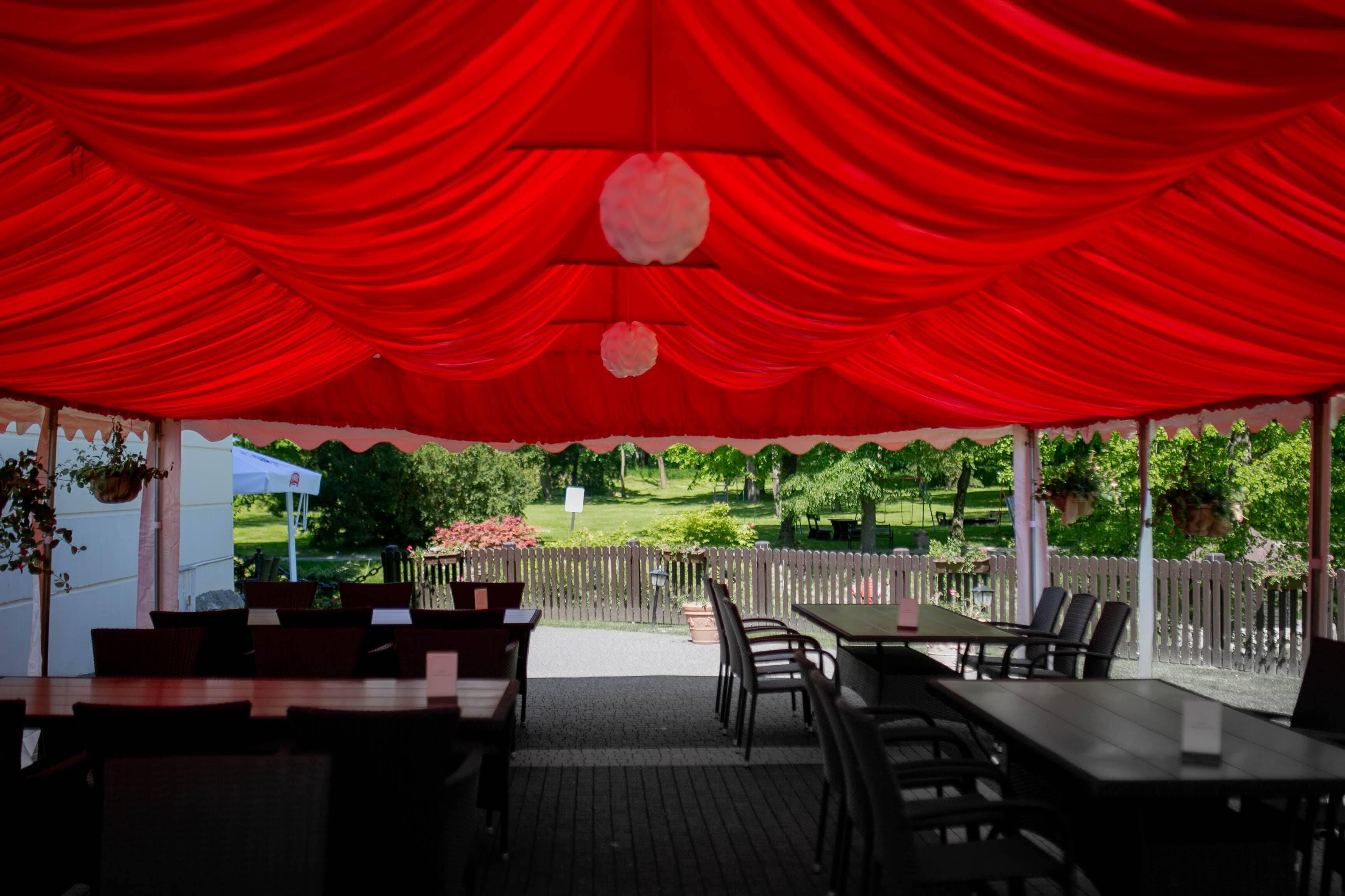 An outdoor covered patio with tables and chairs, a wooden fence, and a view of a garden.
