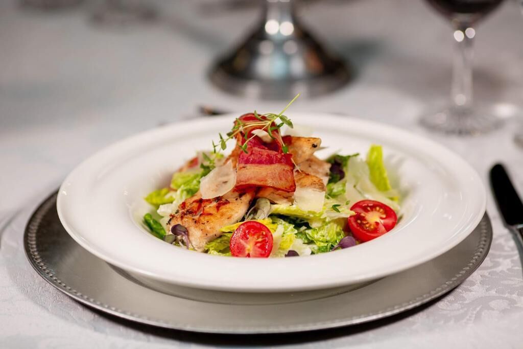 Un plato blanco con ensalada, que incluye lechuga, tomates cherry y pollo a la parrilla, sobre una mesa con un plato base plateado.