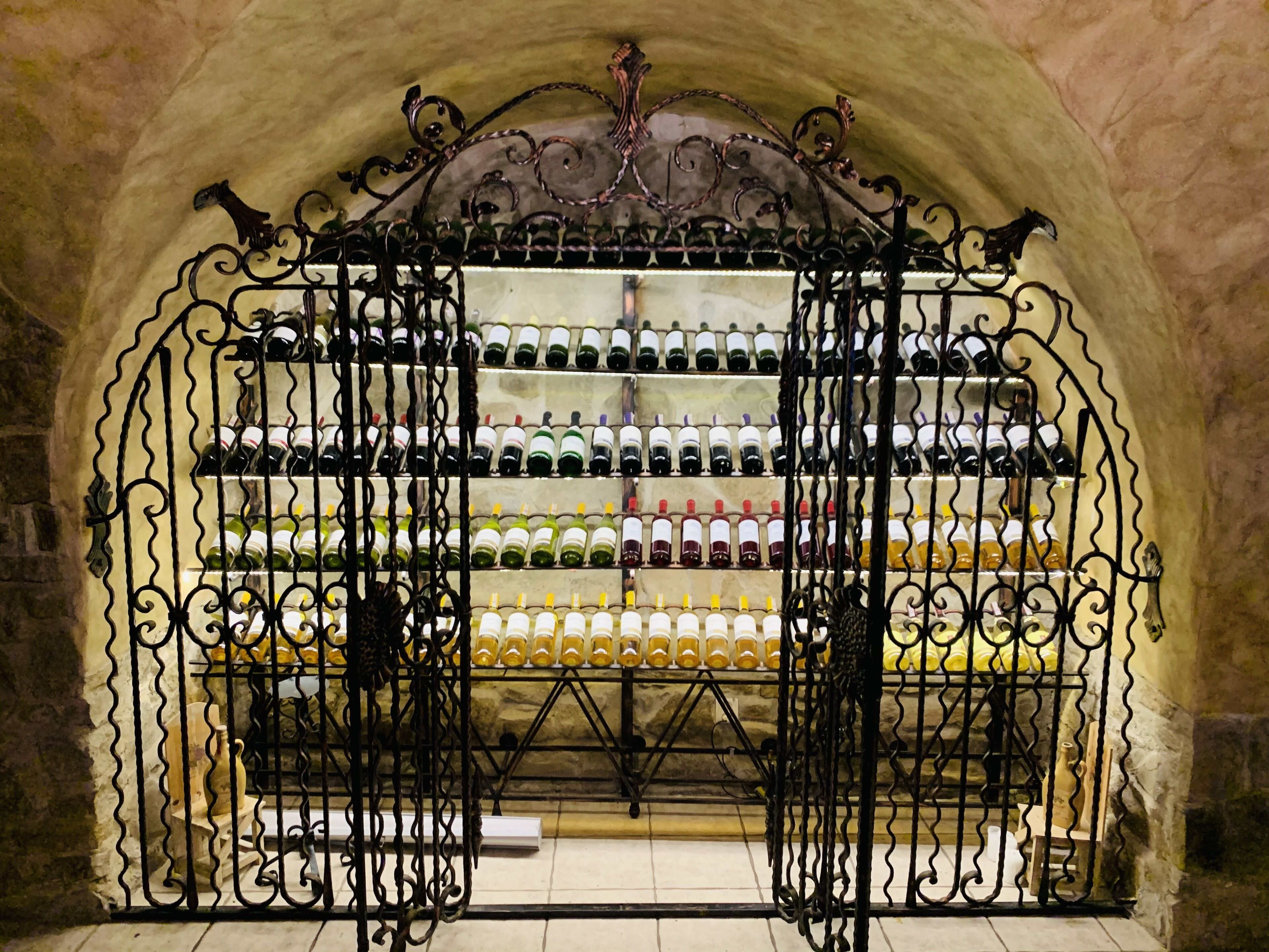 Una bodega abovedada con una reja de hierro forjado, estantes llenos de botellas de vino y paredes de piedra.