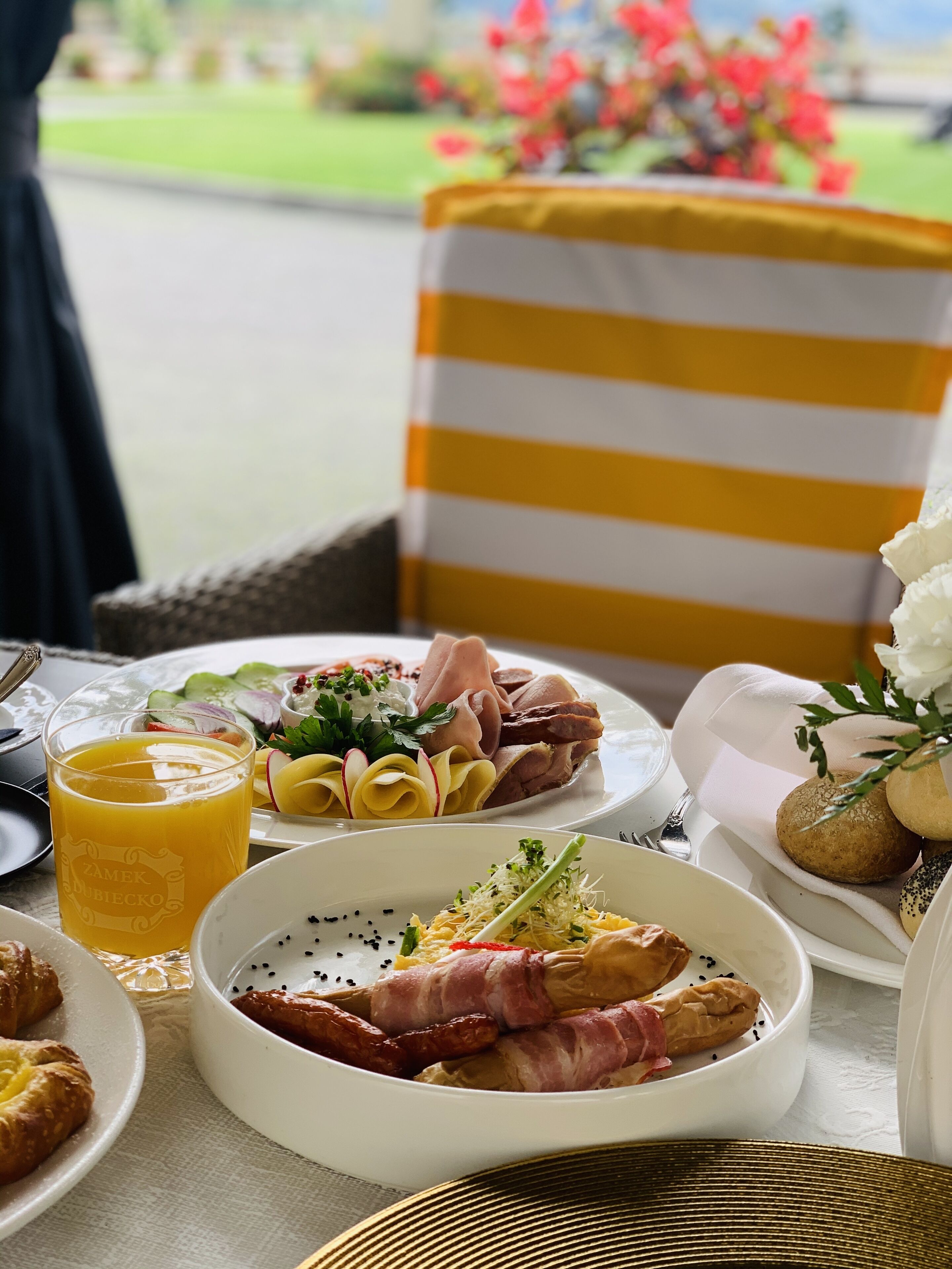 Una mesa servida con diversos platos, entre ellos un vaso de zumo de naranja, una ensalada y platos de carne, con vistas a un jardín al fondo.