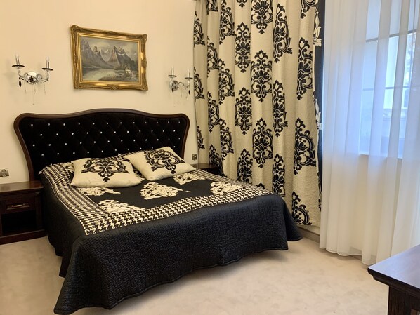 A bedroom with a large bed, a nightstand, a wall-mounted lamp, a framed picture, and patterned curtains.