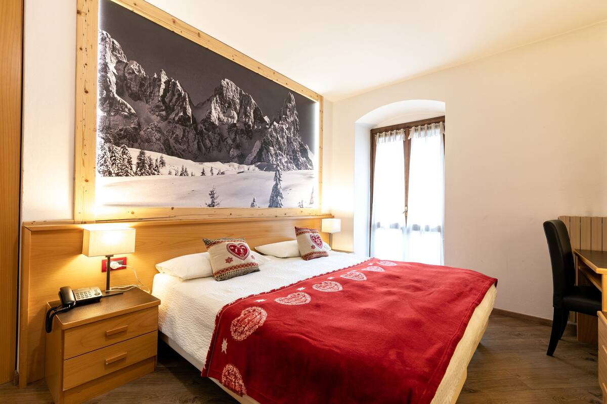A hotel room with a bed, a nightstand with a phone, a desk, and a large mountain painting on the wall.