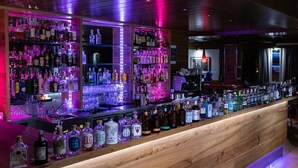 A bar with shelves of liquor bottles and glasses, wooden counter, and purple lighting. - Hotel Isolabella Wellness (Primiero San Martino di Castrozza)