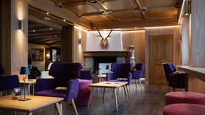 A modern lounge area with purple upholstered chairs, wooden tables, and a stone fireplace. - Hotel Isolabella Wellness (Primiero San Martino di Castrozza)
