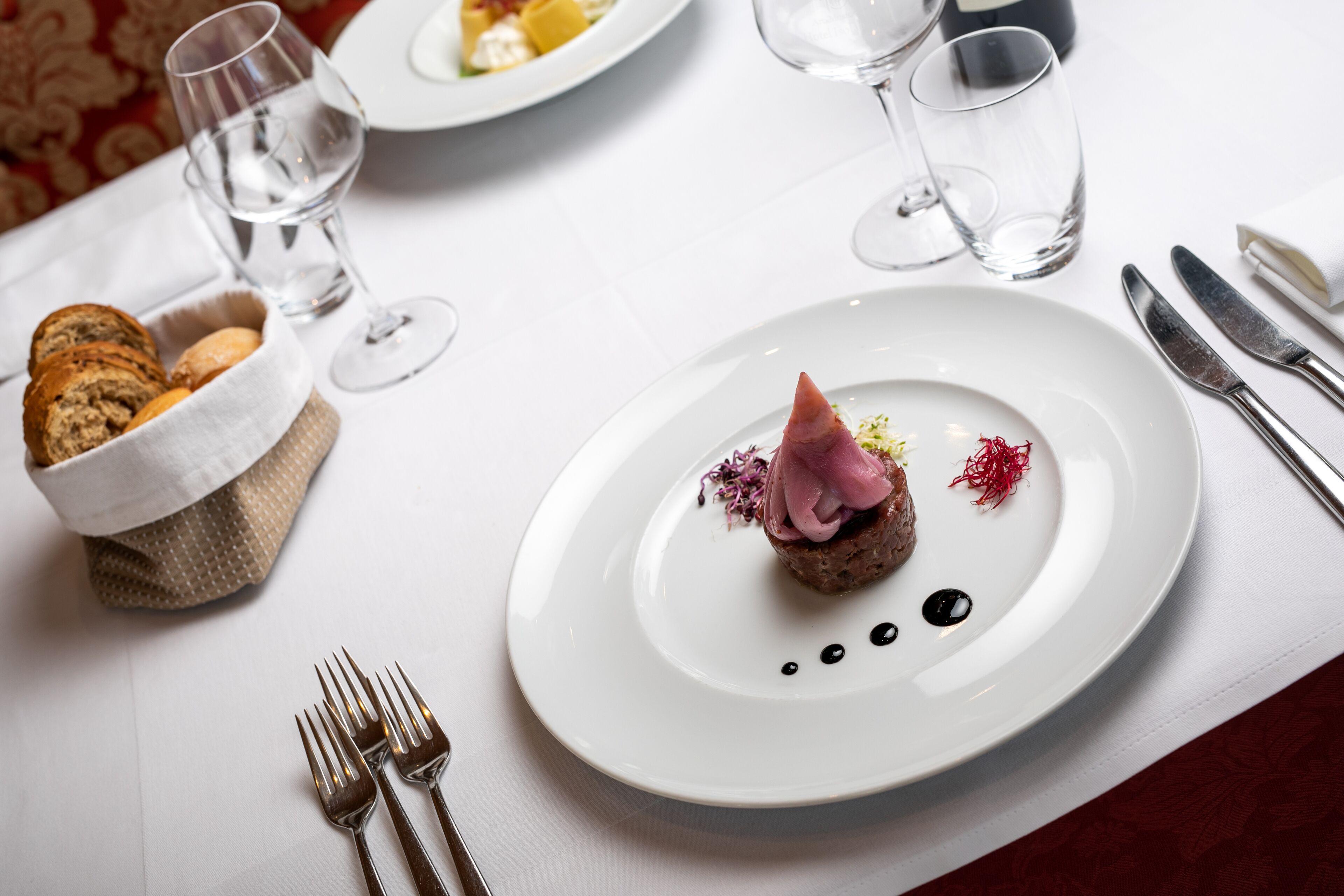 Une table à manger avec une assiette blanche, deux verres à vin, un panier de pain et des couverts.