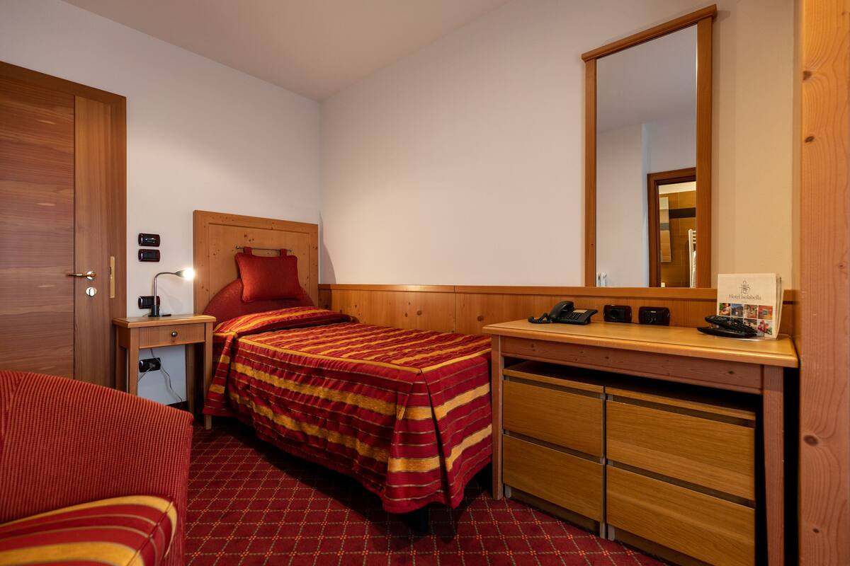 A hotel room with a single bed, a wooden headboard, a desk with a telephone, a mirror, and a small nightstand with a lamp.