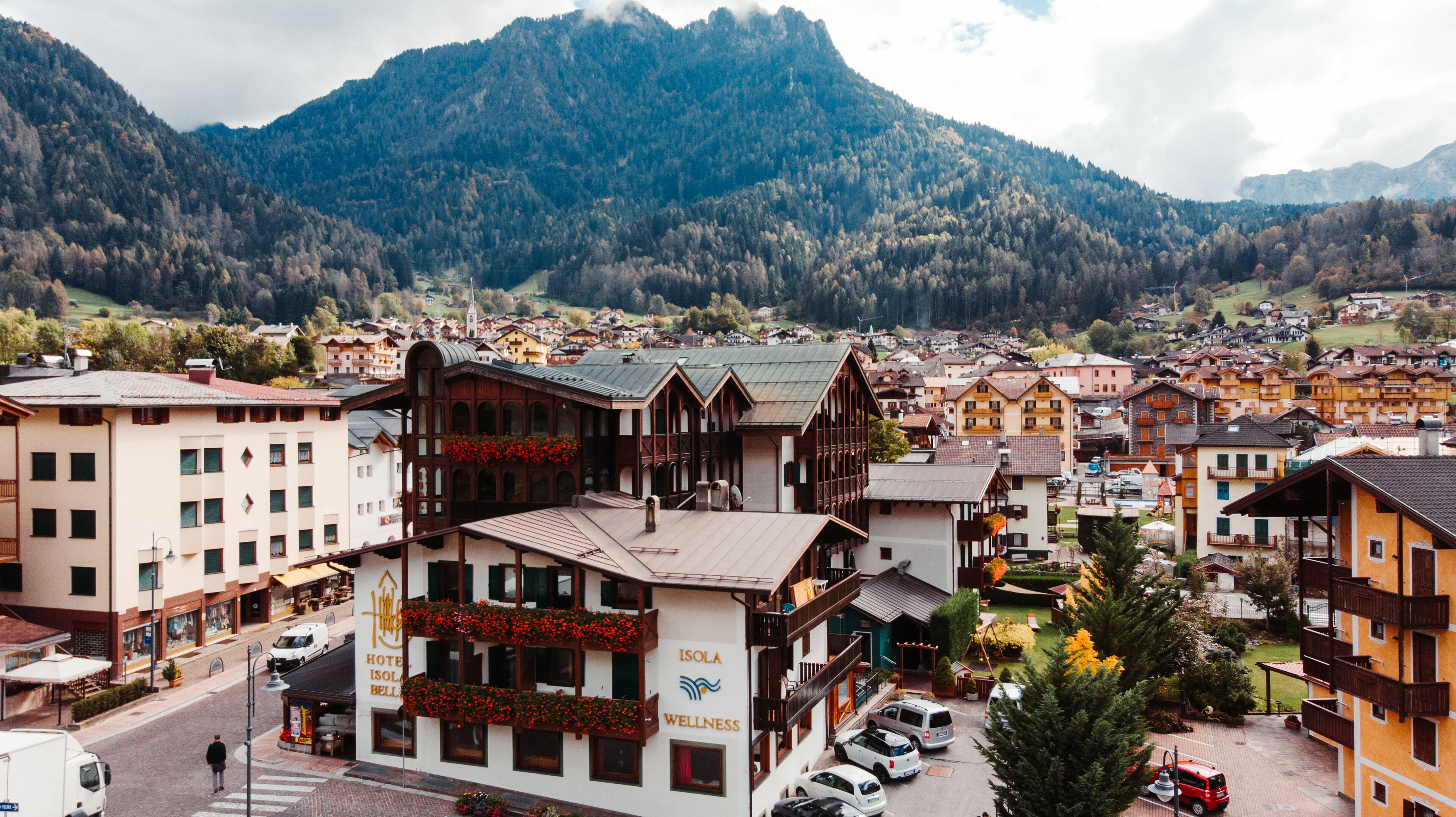 Un hôtel de style alpin traditionnel, doté d’un balcon orné de fleurs rouges, d’une enseigne indiquant « Isola Wellness » et offrant une vue sur le paysage montagneux environnant.