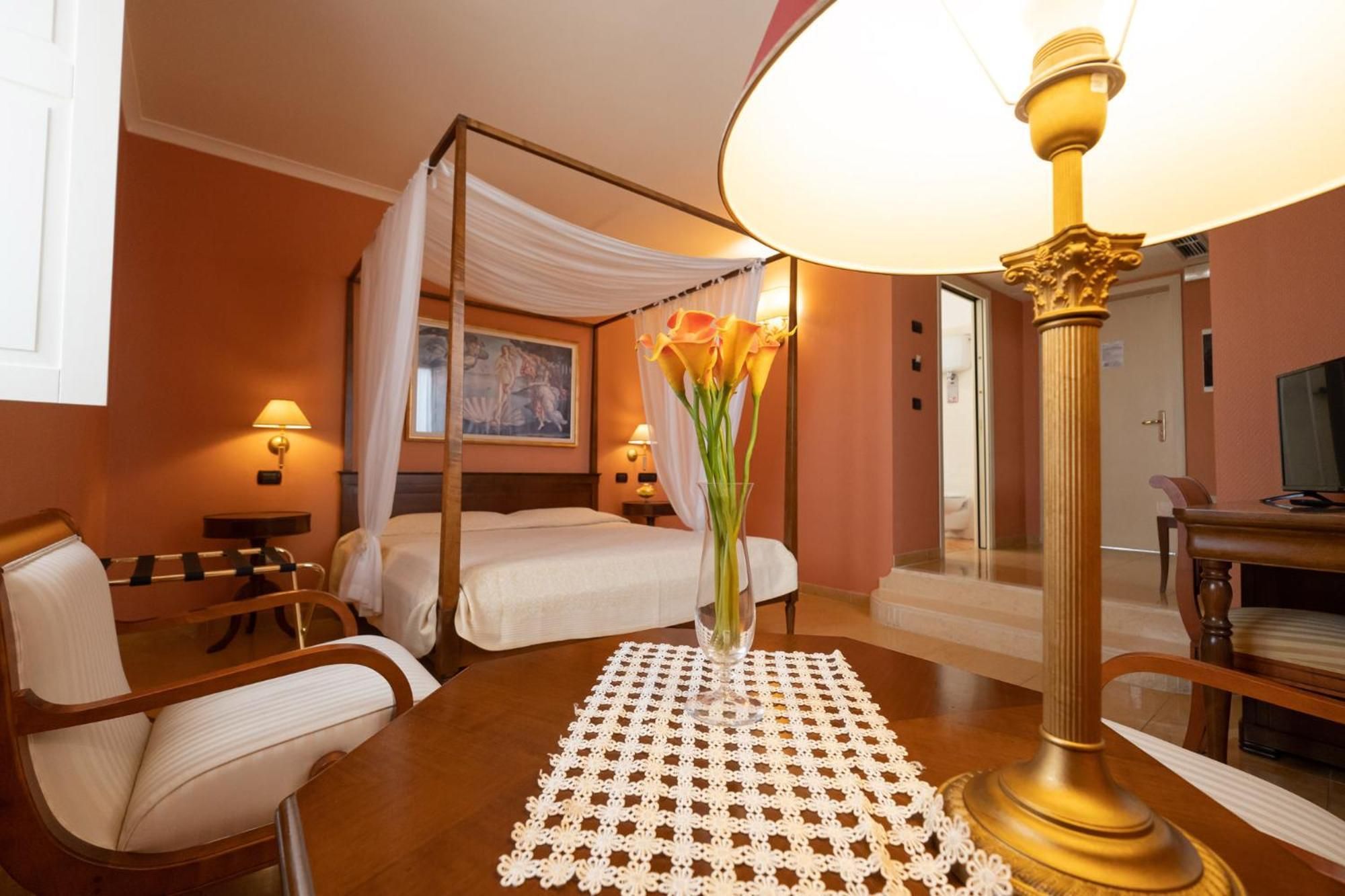 Una camera d'albergo con un letto a baldacchino, un angolo salotto con tavolo e sedie, e un'ampia lampada da terra.