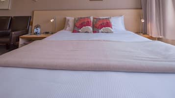 A large bed with a white comforter and two decorative pillows, a bedside table with a lamp, and a chair in a hotel room.