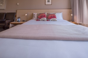 A large bed with a white comforter and two decorative pillows, a bedside table with a lamp, and a chair in a hotel room. - Amross Court Motor Lodge (Christchurch)