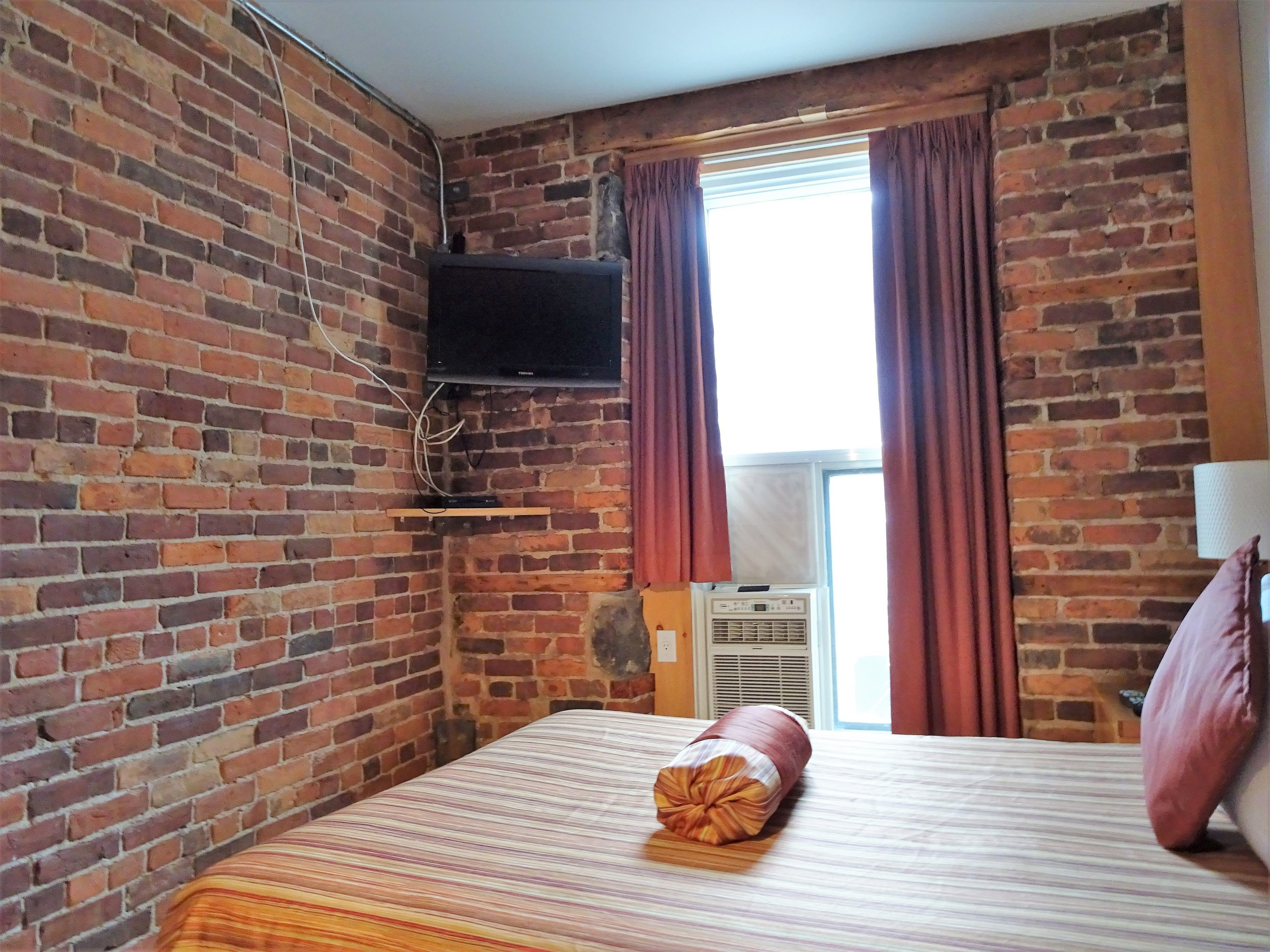 Habitación de hotel con pared de ladrillo, televisor de pantalla plana montado en la pared, cama con colcha a rayas y ventana con cortinas.