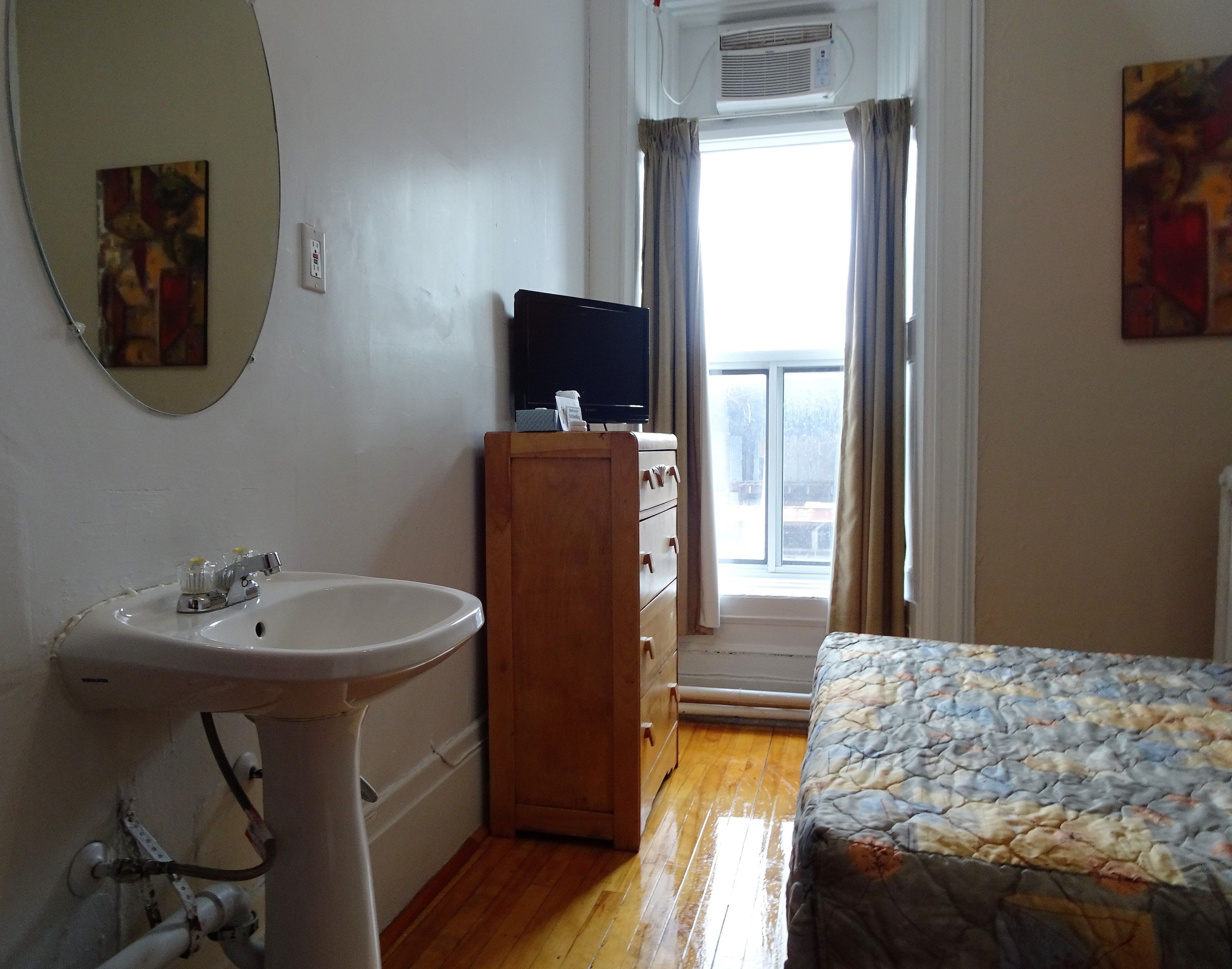 Una habitación con un lavamanos blanco, cómoda de madera, un televisor de pantalla plana encima y una cama con una colcha estampada, además de una ventana con cortinas.