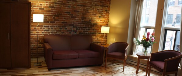 A room with a brick wall, a wooden floor, a red sofa, two wooden chairs, a small table, and a window with a view of buildings.