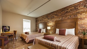 A hotel room with two beds, a wooden desk, a television, and a brick wall.