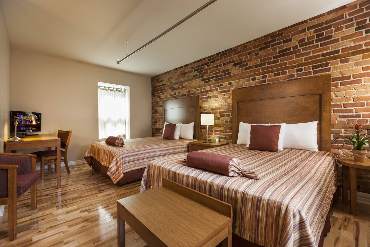 A hotel room with two beds, a wooden desk, a television, and a brick wall.