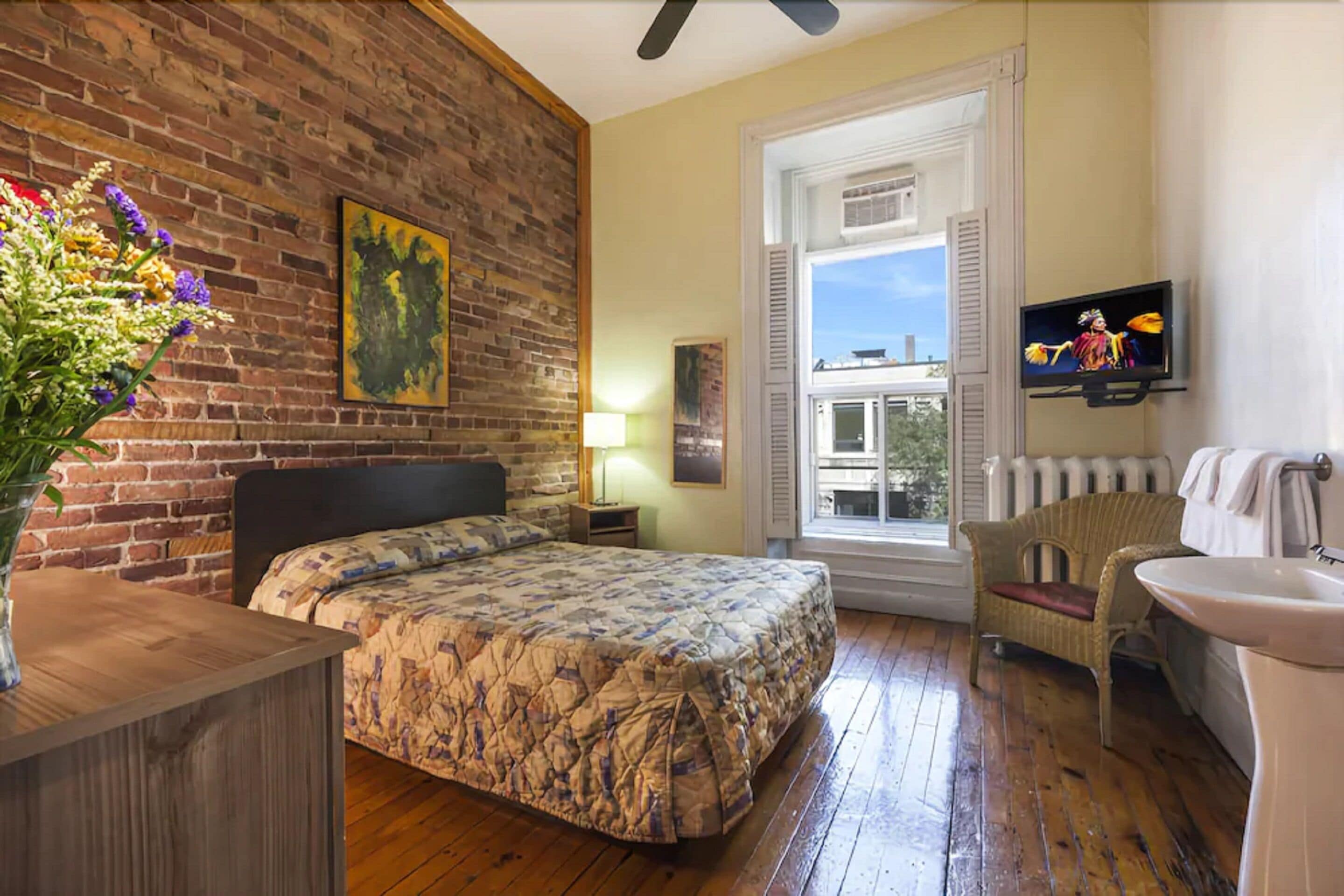 A bedroom with a brick wall, a bed with a floral bedspread, a window with shutters, a wicker chair, a TV, and a radiator.