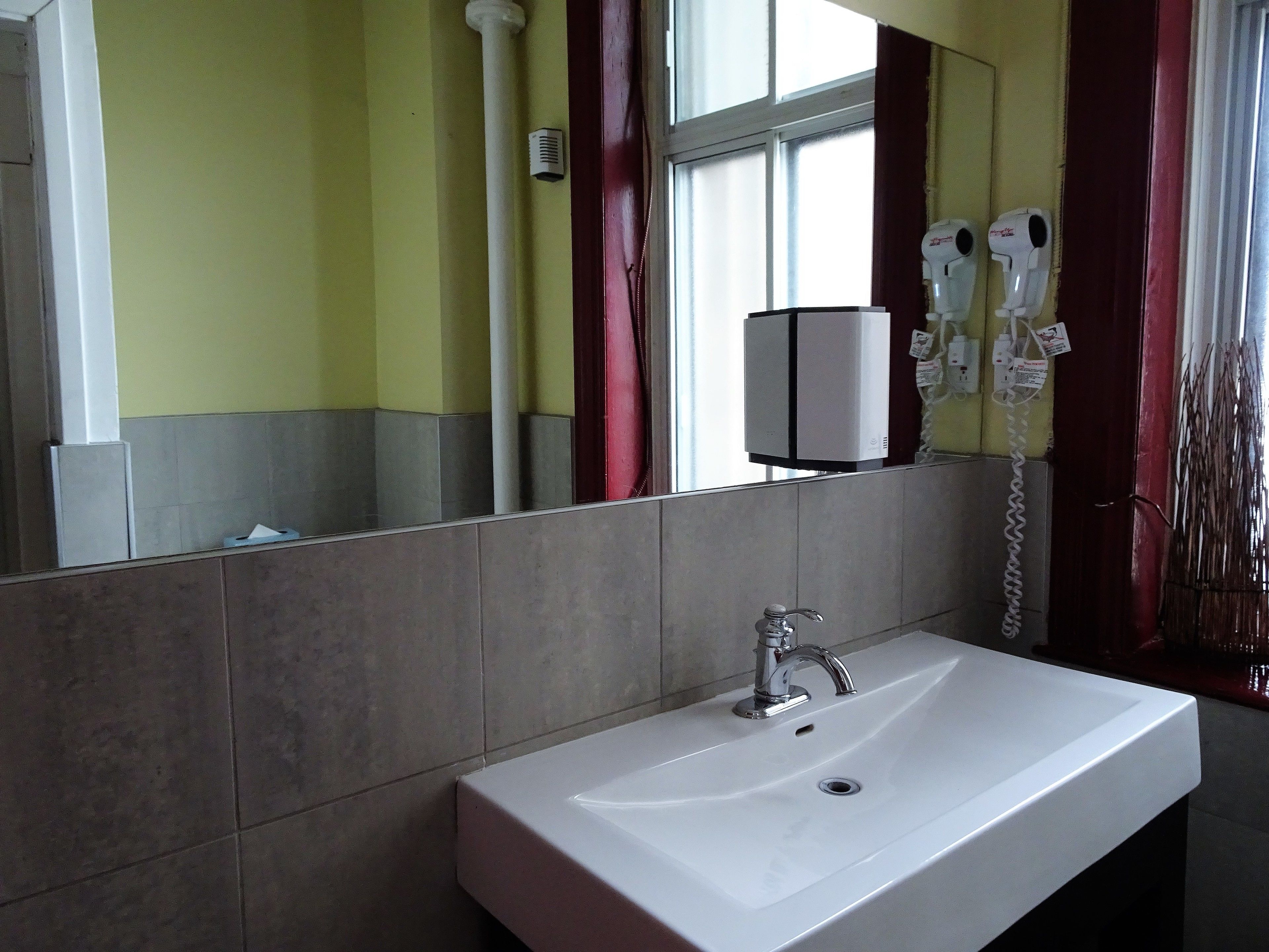 Un baño con lavamanos blanco, un espejo, una ventana con cortinas rojas y dos secadores de pelo fijados en la pared.