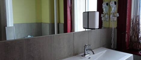 Un baño con lavamanos blanco, un espejo, una ventana con cortinas rojas y dos secadores de pelo fijados en la pared.