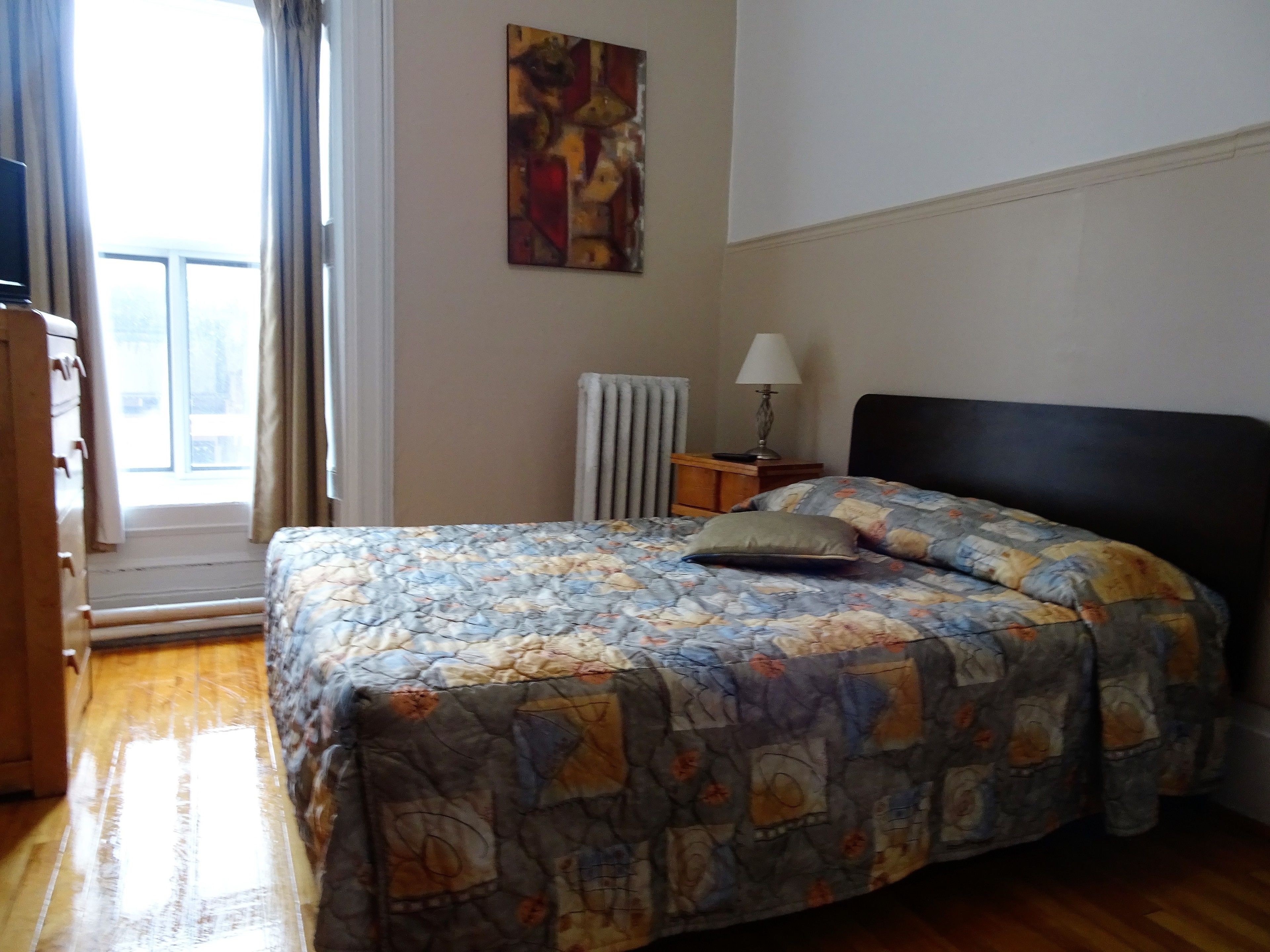 Un dormitorio con cama, mesita de noche, lámpara, radiador y ventana con cortinas.