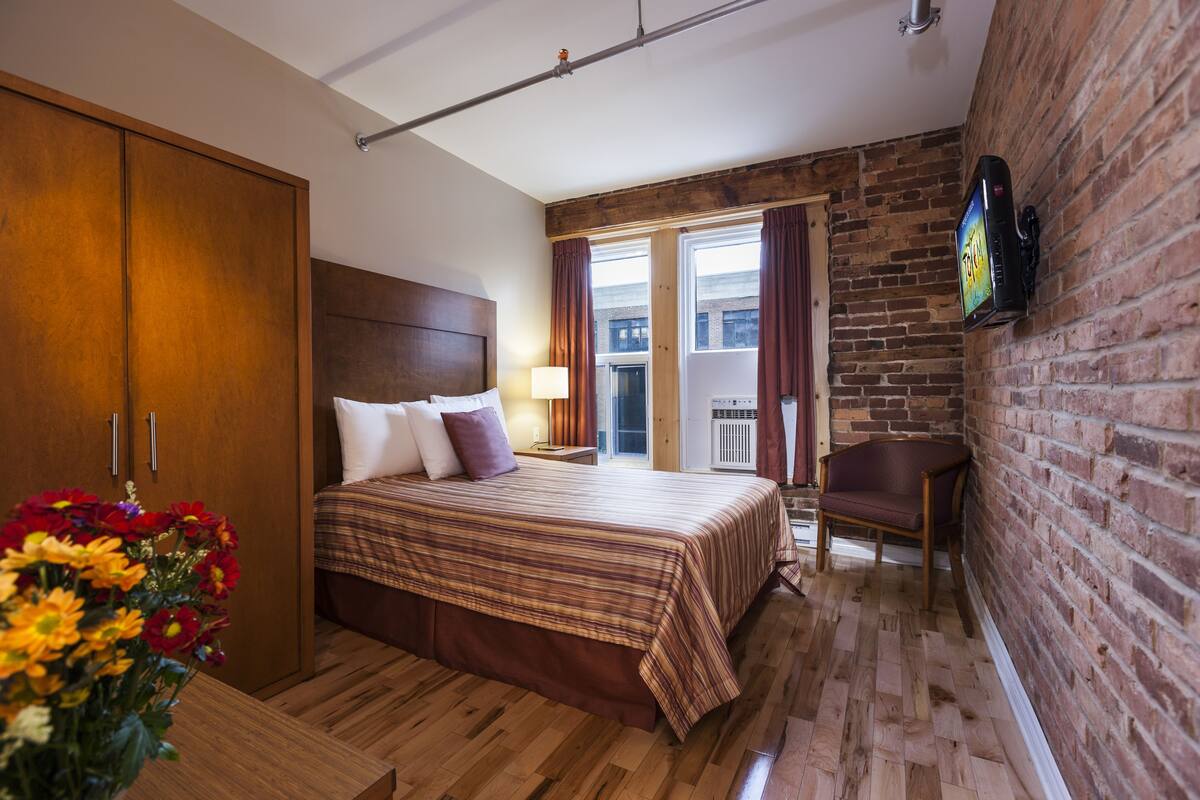 A hotel room with a wooden floor, a bed with a striped bedspread, a wooden wardrobe, a chair, and a TV mounted on the brick wall.