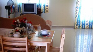 A dining area with a wooden table set for two, a television in the background, and a sofa. - VillaOSoleil Apartments (Grand-Baie)
