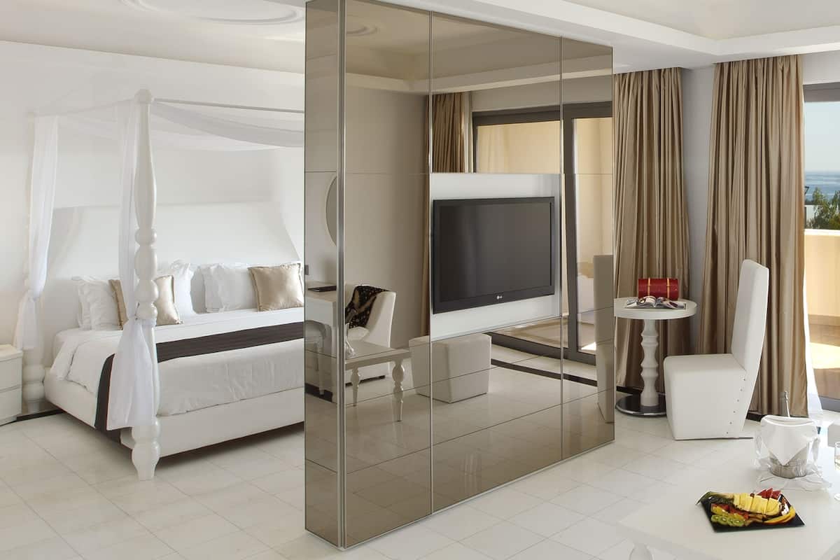 Quarto de hotel moderno com cama de dossel, televisão, área de jantar com mesa e cadeiras e varanda com cortinas.