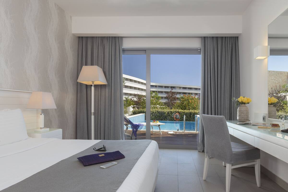 Quarto de hotel com cama, escrivaninha, cadeira, luminária e vista para uma piscina e um edifício.