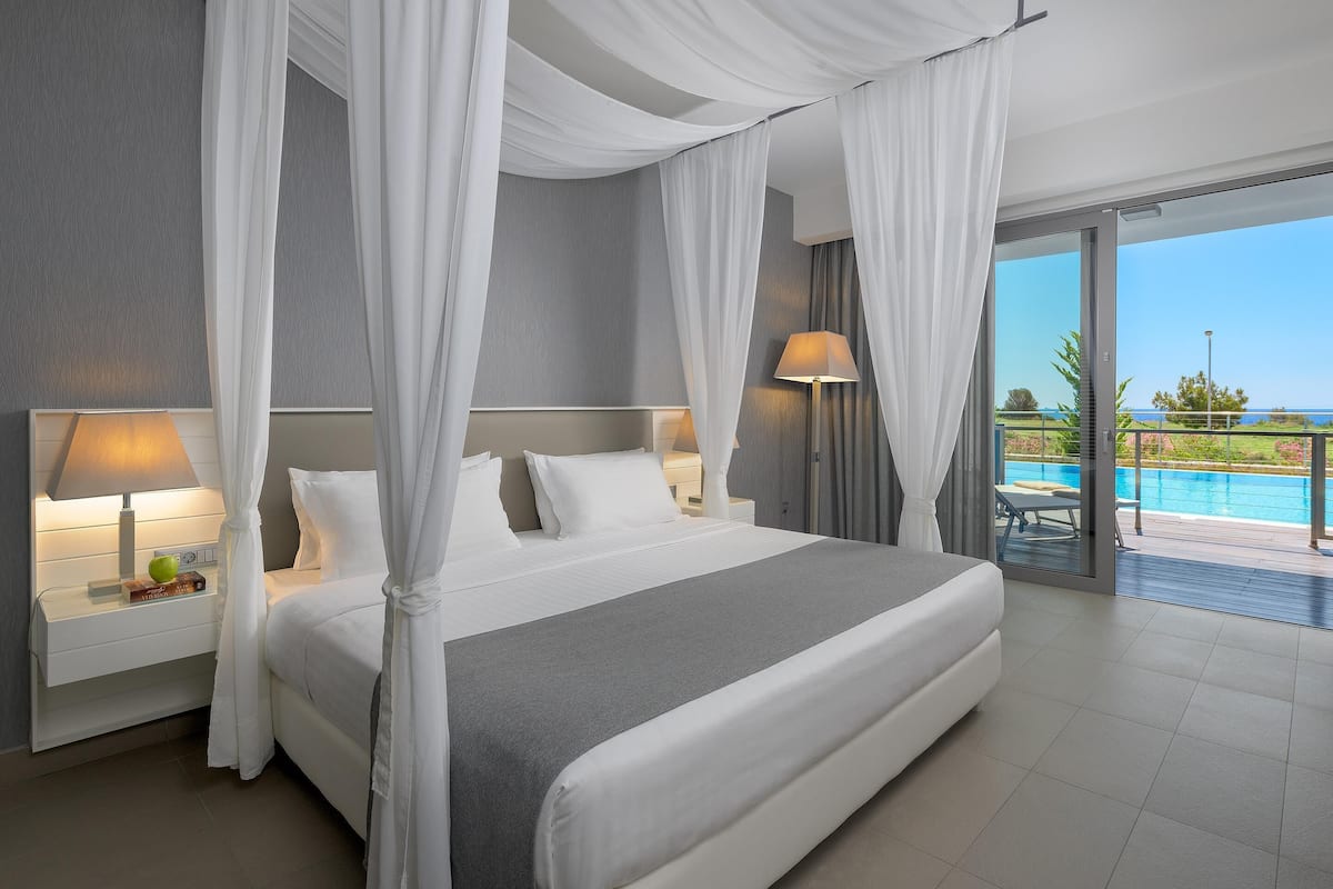 Quarto moderno com uma cama grande, cortinas brancas, mesa de cabeceira com luminária e vista para uma piscina.