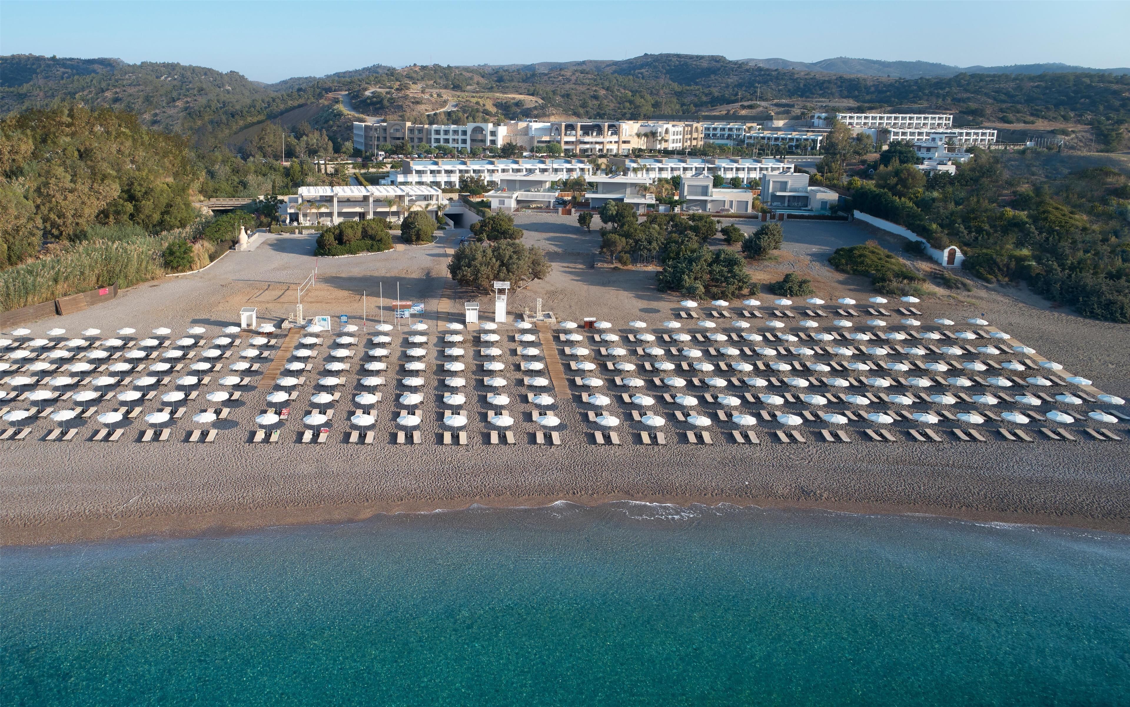 Vista aérea de un complejo turístico frente a la playa, con hileras de sombrillas y tumbonas, un aparcamiento y un conjunto de edificios al fondo.