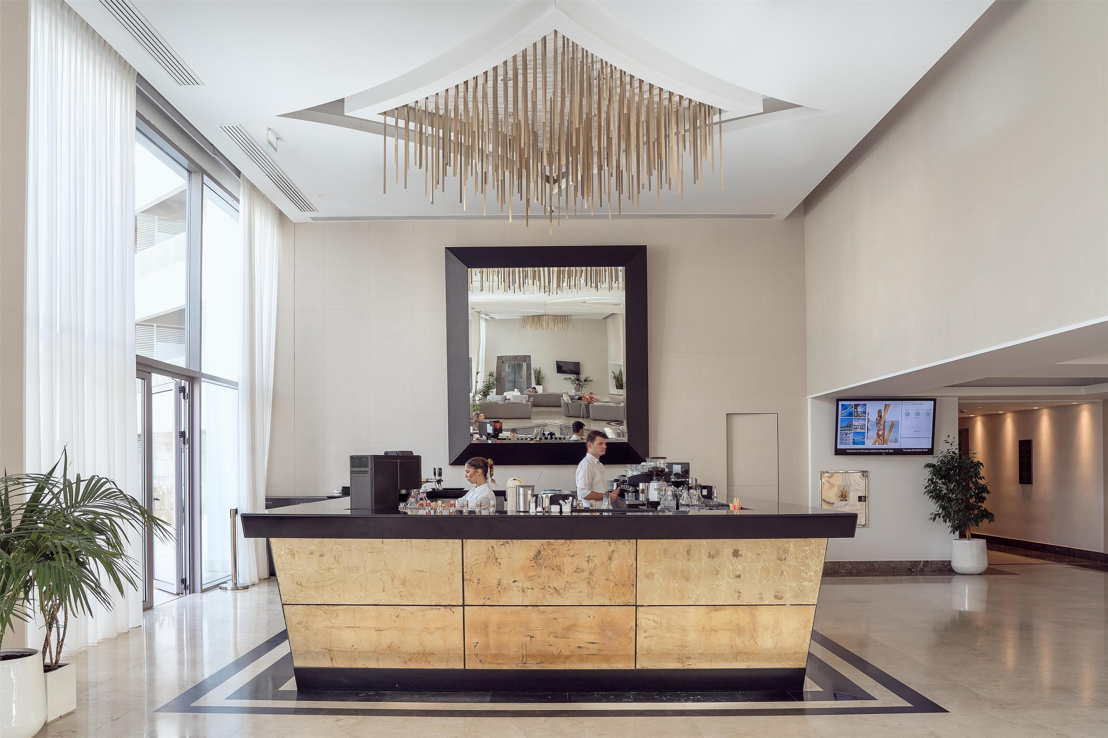 A modern hotel lobby with a reception desk, large mirror, and a television screen.