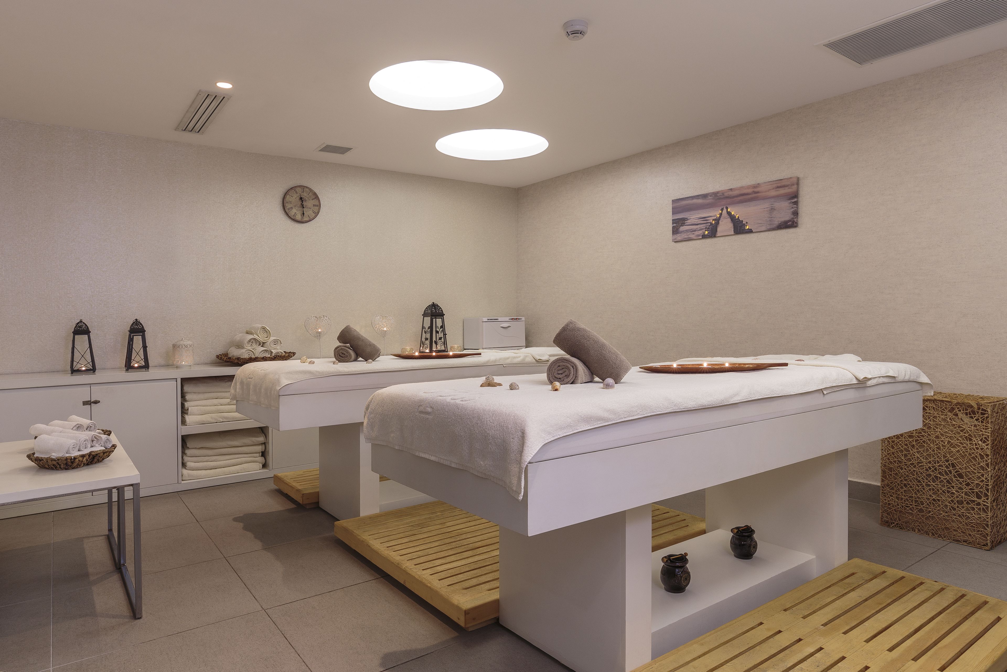 Une salle de spa équipée de deux tables de massage, d’une petite table avec des serviettes, d’une horloge et d’un tableau au mur.