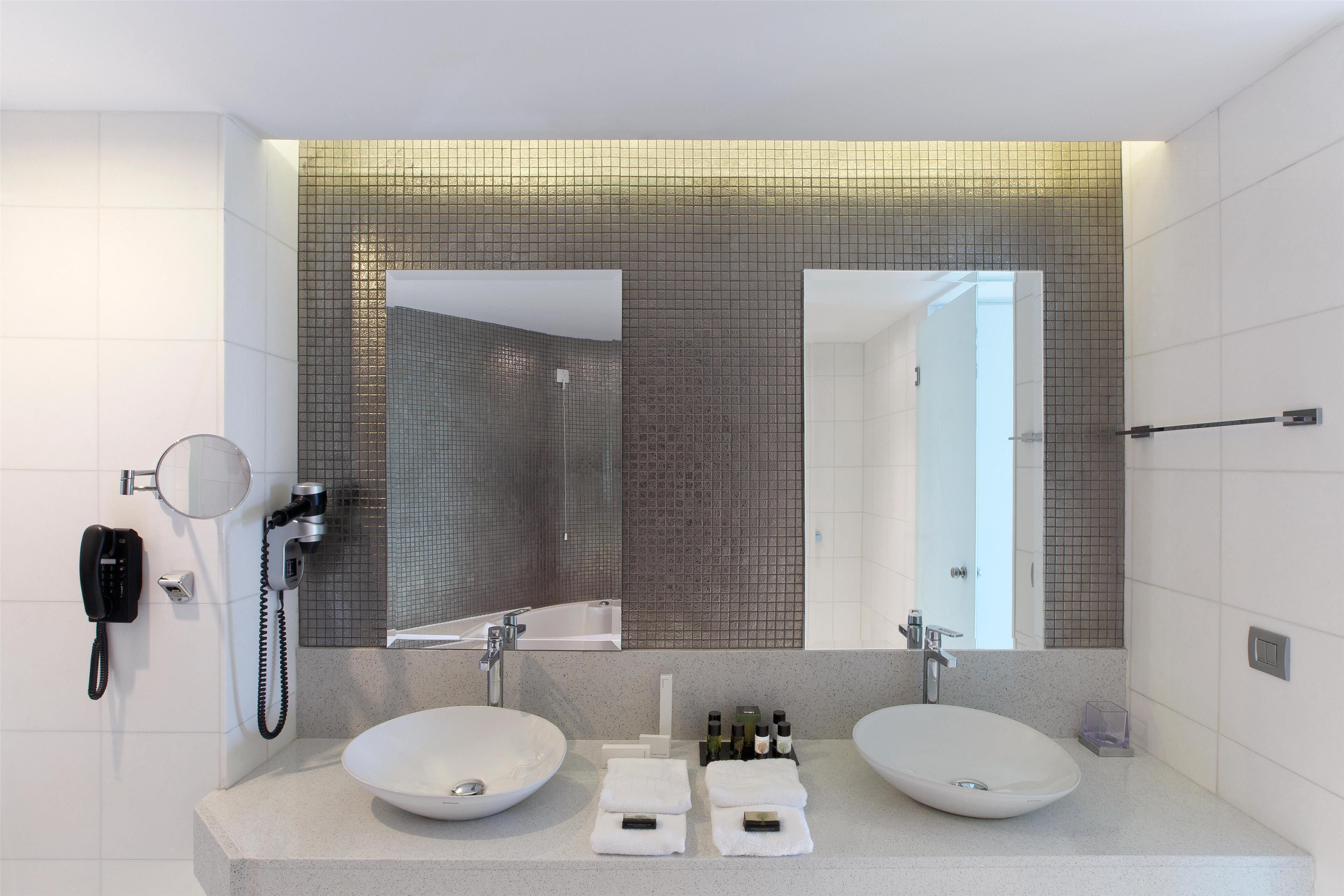 A modern bathroom with two sinks, a large mirror, a handheld showerhead, and a telephone.