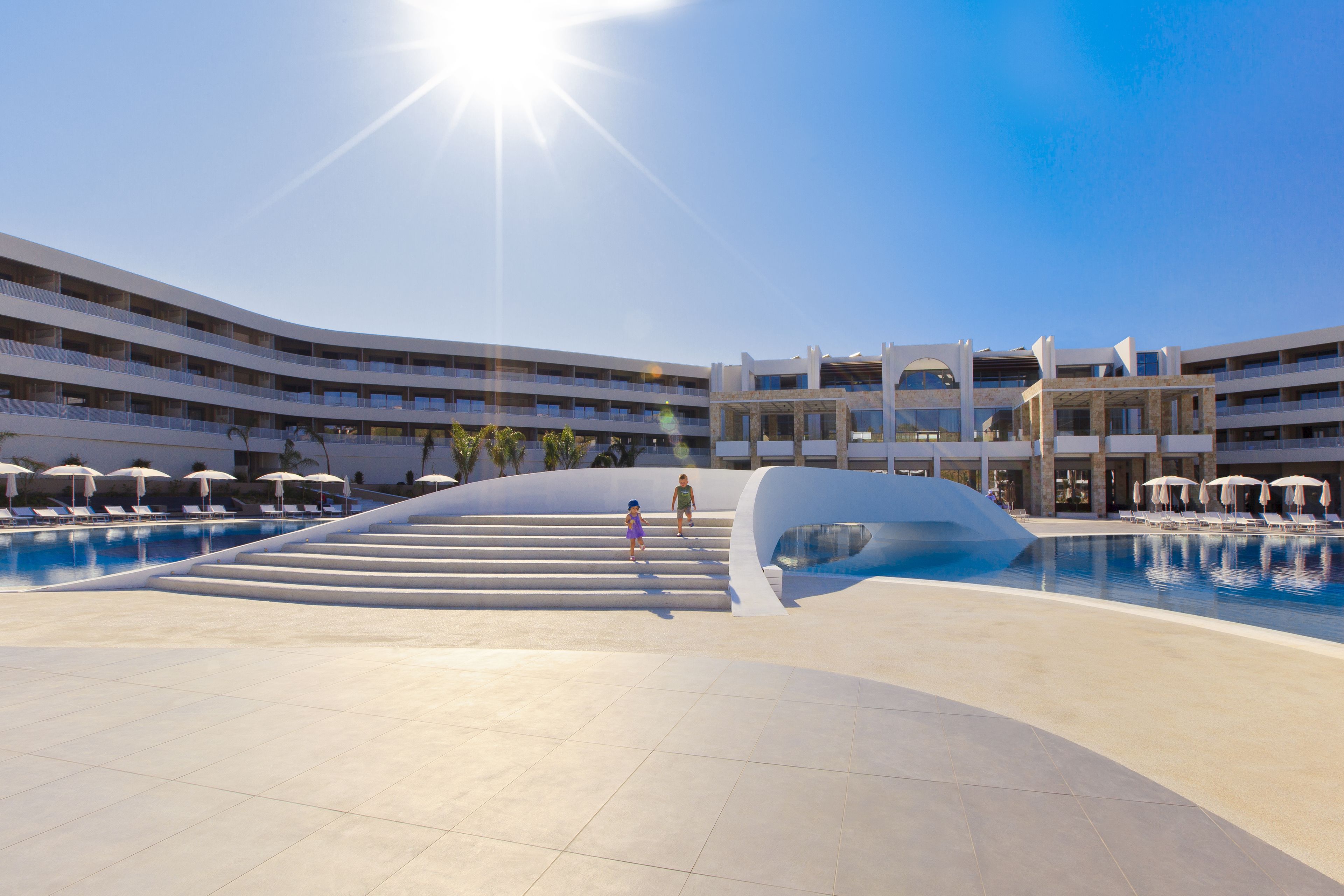 A modern hotel with a central pool, multiple balconies, and a sunlit sky.