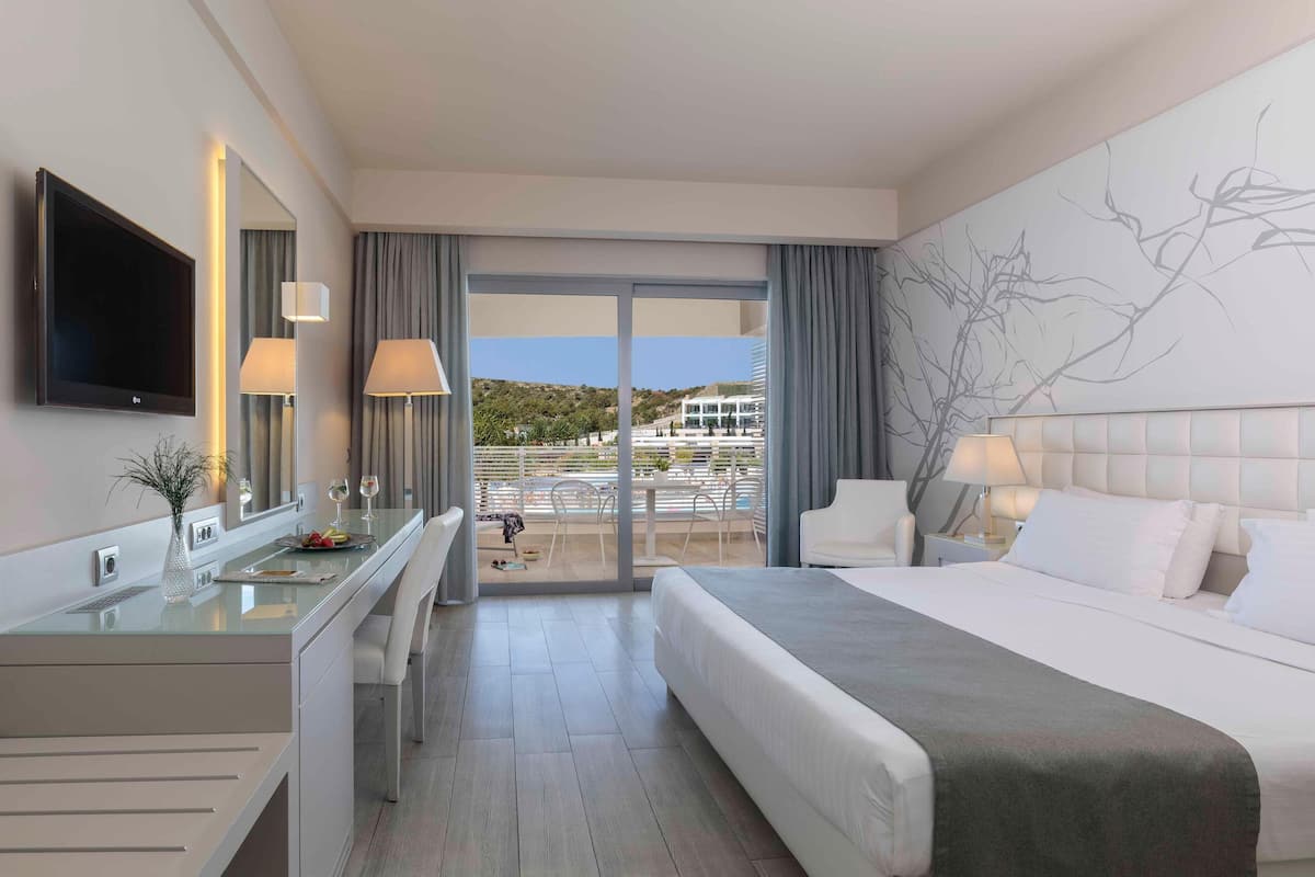 Quarto de hotel moderno com uma cama grande, uma escrivaninha com televisão de tela plana, uma varanda com vista para um edifício e árvores, e uma janela ampla com cortinas.
