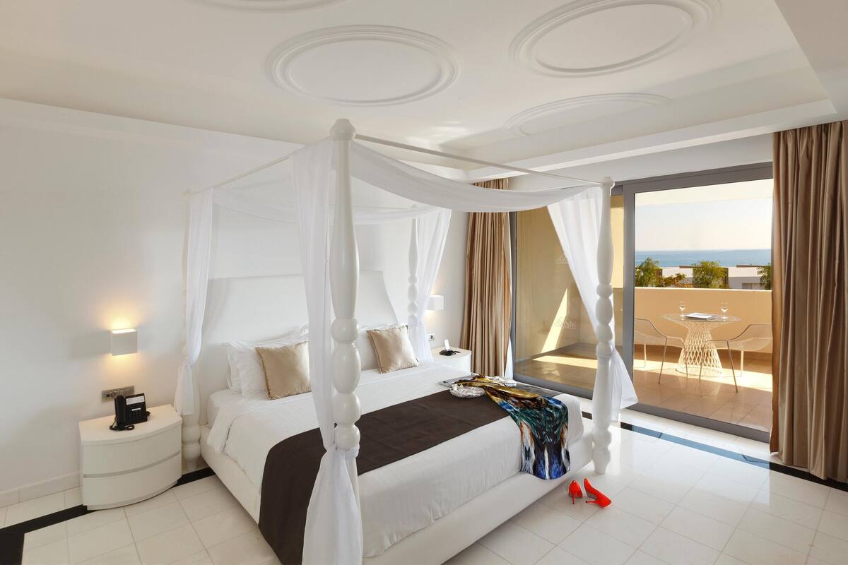 Quarto com cama de dossel, mesa de cabeceira com telefone e vista para uma varanda com mesa e cadeiras.