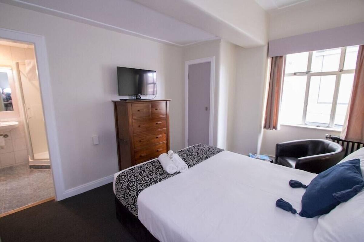 Habitación de hotel con una cama, un televisor en una cómoda, una silla y un ventanal con cortinas.