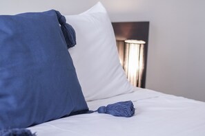 A bed with white sheets and blue pillows, a blue tassel, and a modern bedside lamp.