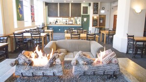 L’intérieur d’un restaurant présente un espace central avec des sièges, notamment un foyer, plusieurs tables et chaises, ainsi qu’un comptoir derrière lequel se trouve une personne.