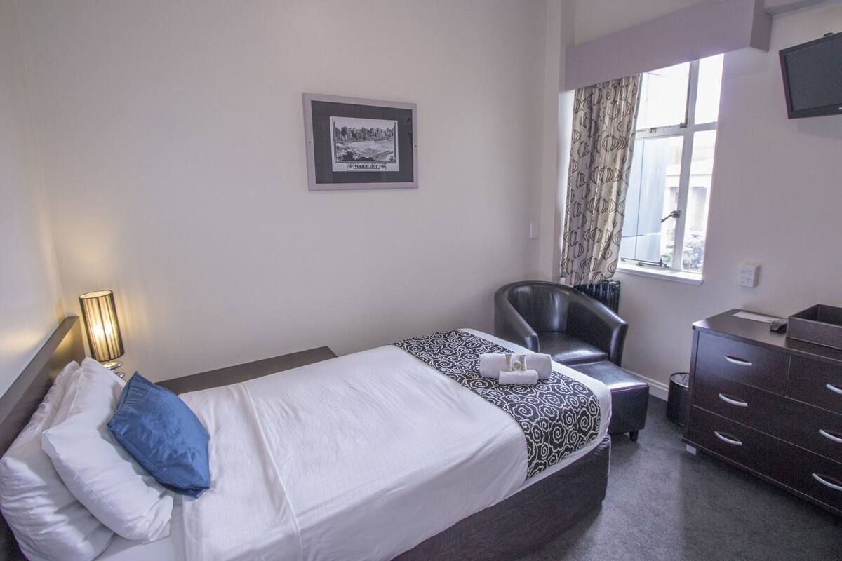 Habitación de hotel con cama, una silla, cómoda, lámpara, ventana con cortinas e imagen enmarcada en la pared.