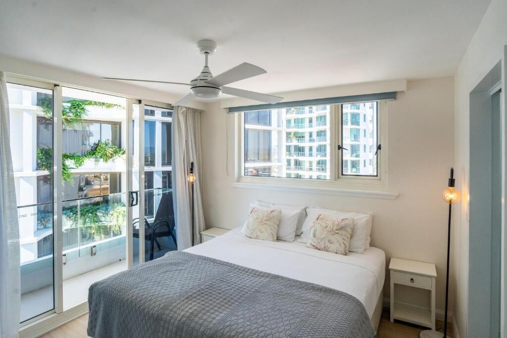 A bedroom with a bed, a ceiling fan, a nightstand, a floor lamp, and a view of the outside.